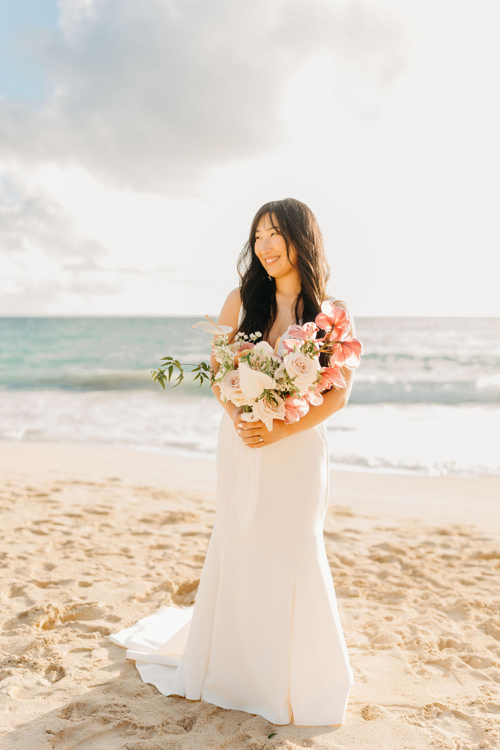A couple in wedding attire embraces on a beach, with the ocean and sky in the background. The woman holds a bouquet and both appear happy for their Waimanalo Beach elopement 