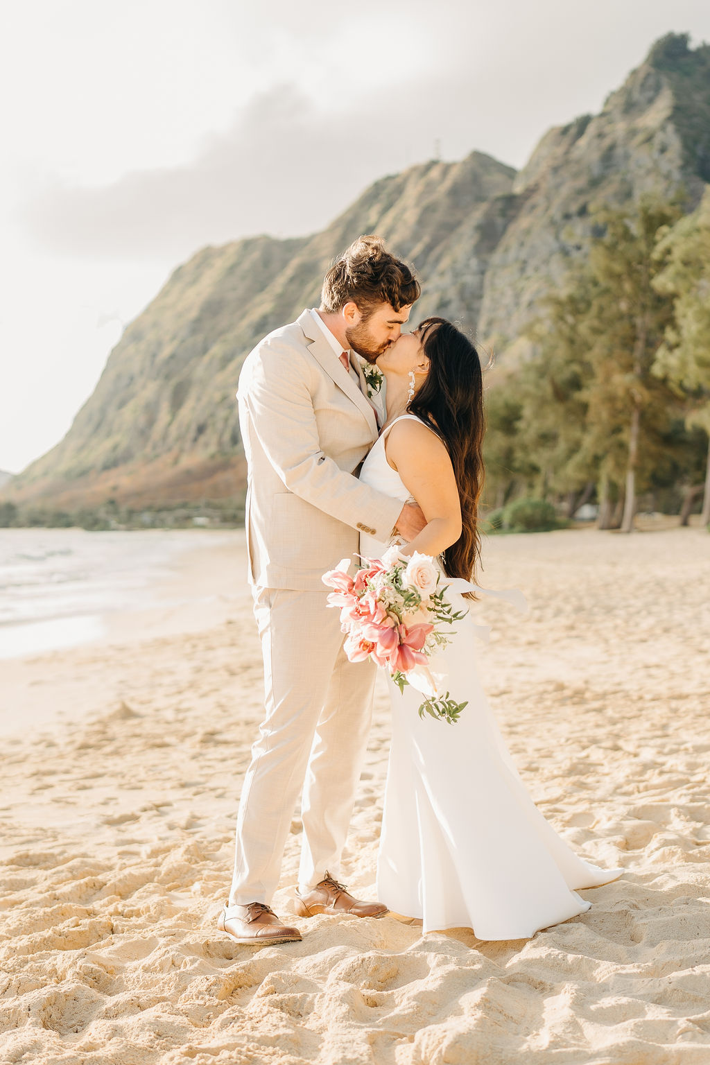 A couple in wedding attire embraces on a beach, with the ocean and sky in the background. The woman holds a bouquet and both appear happy for their Waimanalo Beach elopement 