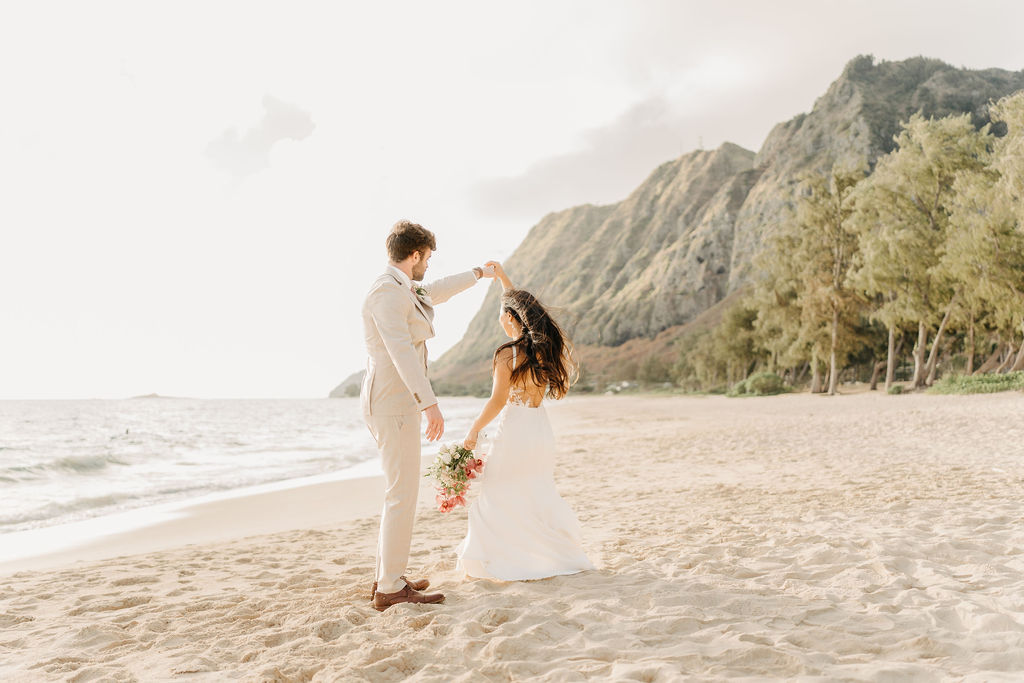 A couple in wedding attire embraces on a beach, with the ocean and sky in the background. The woman holds a bouquet and both appear happy for their Waimanalo Beach elopement 