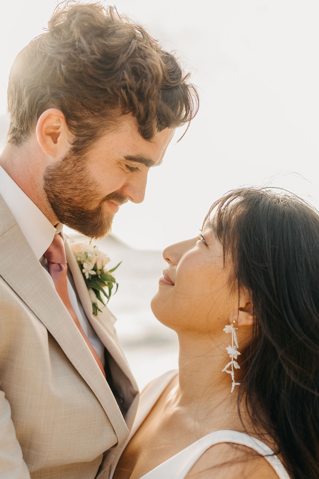 A couple in wedding attire embraces on a beach, with the ocean and sky in the background. The woman holds a bouquet and both appear happy for their Waimanalo Beach elopement