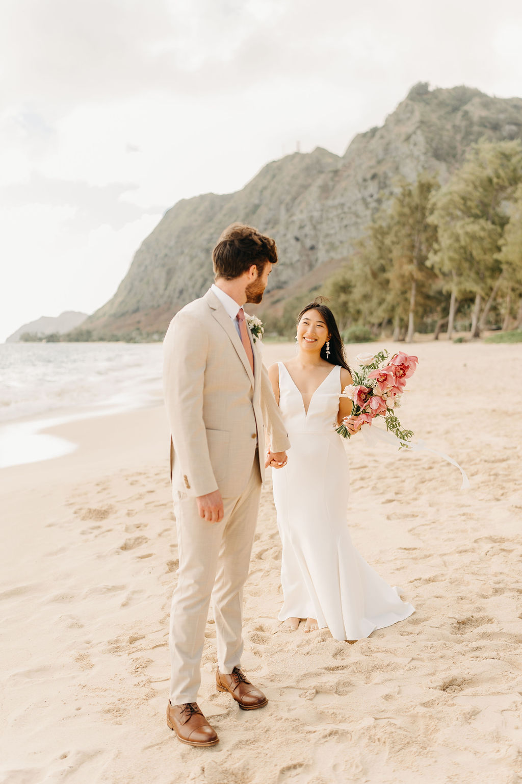 A couple in wedding attire embraces on a beach, with the ocean and sky in the background. The woman holds a bouquet and both appear happy for their Waimanalo Beach elopement