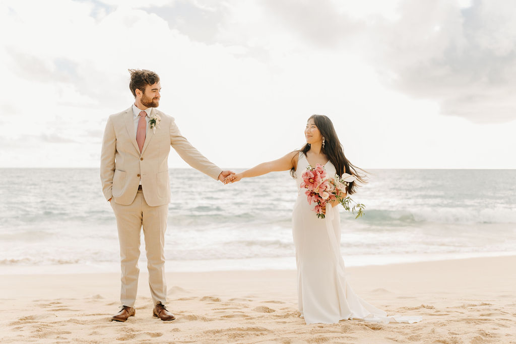 A couple in wedding attire embraces on a beach, with the ocean and sky in the background. The woman holds a bouquet and both appear happy for their Waimanalo Beach elopement