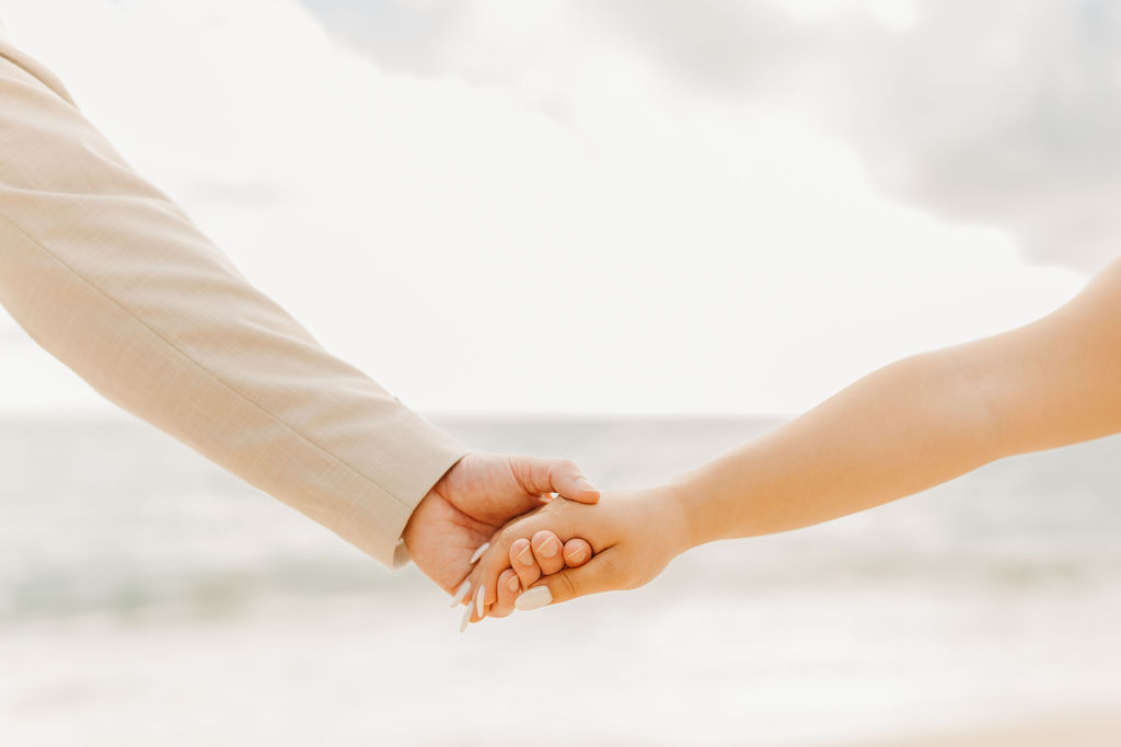 A couple in wedding attire embraces on a beach, with the ocean and sky in the background. The woman holds a bouquet and both appear happy for their Waimanalo Beach elopement