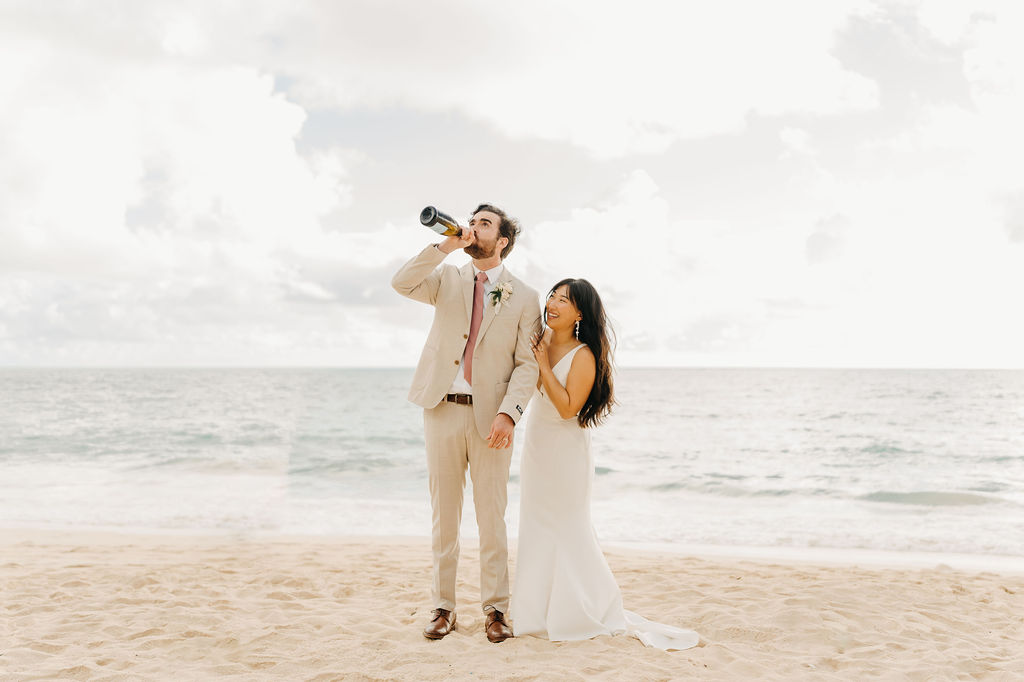 A couple in wedding attire embraces on a beach, with the ocean and sky in the background. The woman holds a bouquet and both appear happy for their Waimanalo Beach elopement
