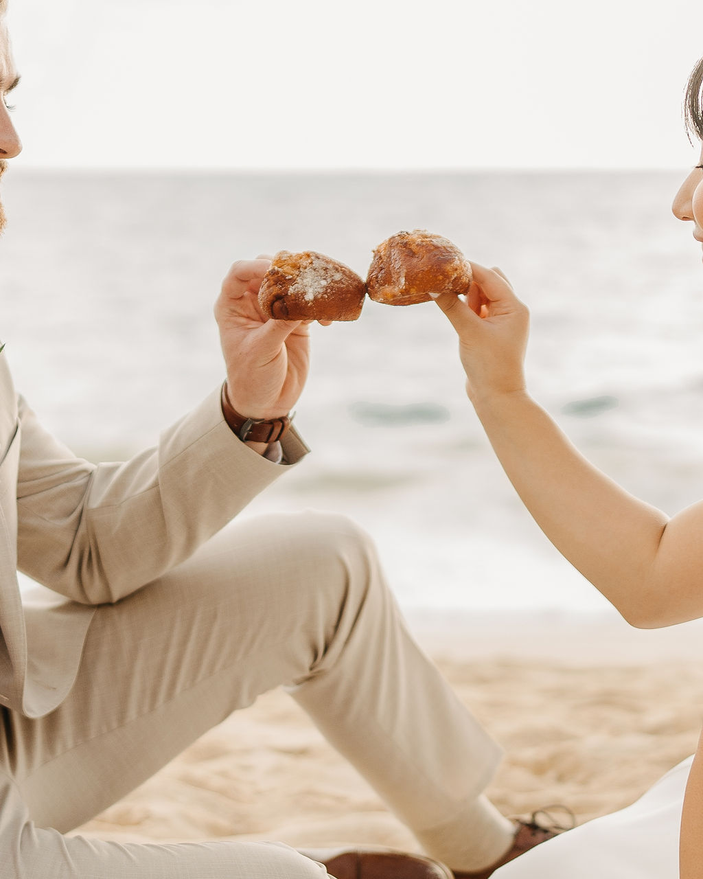A couple in wedding attire embraces on a beach, with the ocean and sky in the background. The woman holds a bouquet and both appear happy for their Waimanalo Beach elopement
