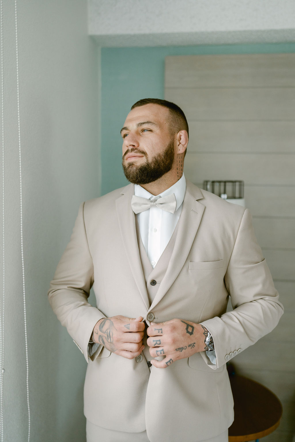 A man with tattoos wears a light beige suit and bow tie, standing indoors and buttoning his jacket while looking to the sidefor his wedding at queen kapiolani