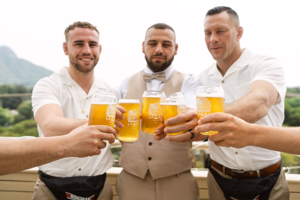 Three men, two in white shirts and one in a beige vest, raise glasses of beer in a toast outdoors with mountains and greenery in the background.
