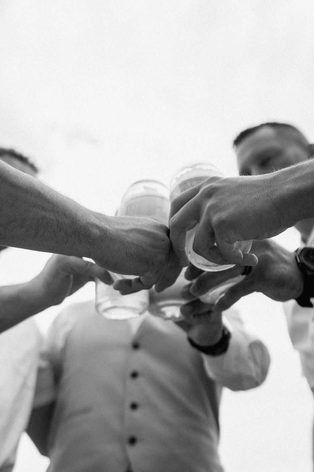 Three men, two in white shirts and one in a beige vest, raise glasses of beer in a toast outdoors with mountains and greenery in the background.