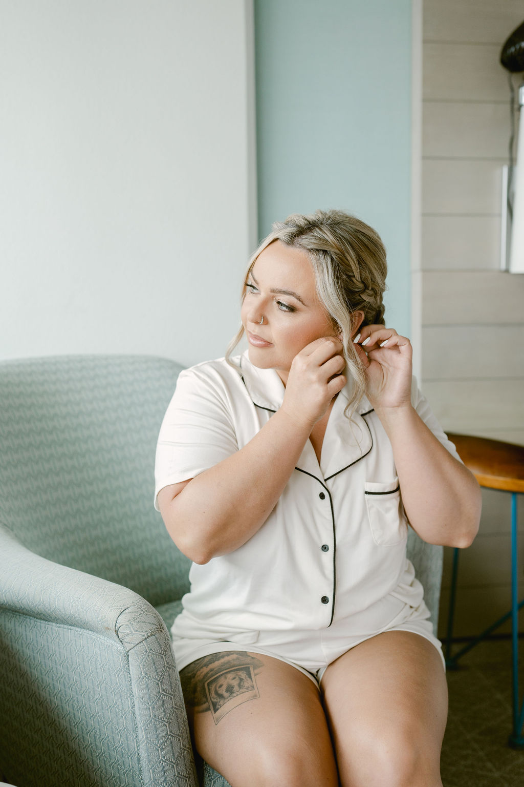 Woman wearing light-colored pajamas sits on a chair and puts on an earring, looking slightly to the side, with a tattoo visible on her thigh.
