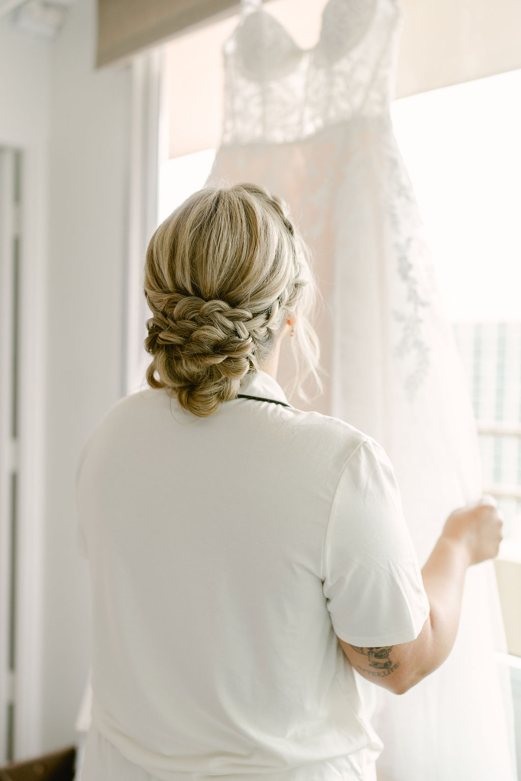 A woman with braided blonde hair, seen from behind, holds up a white lace wedding dress near a window.