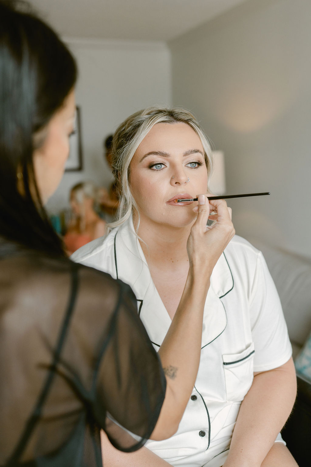 A woman in white pajamas sits while having her makeup applied by a makeup artist in a well-lit room.