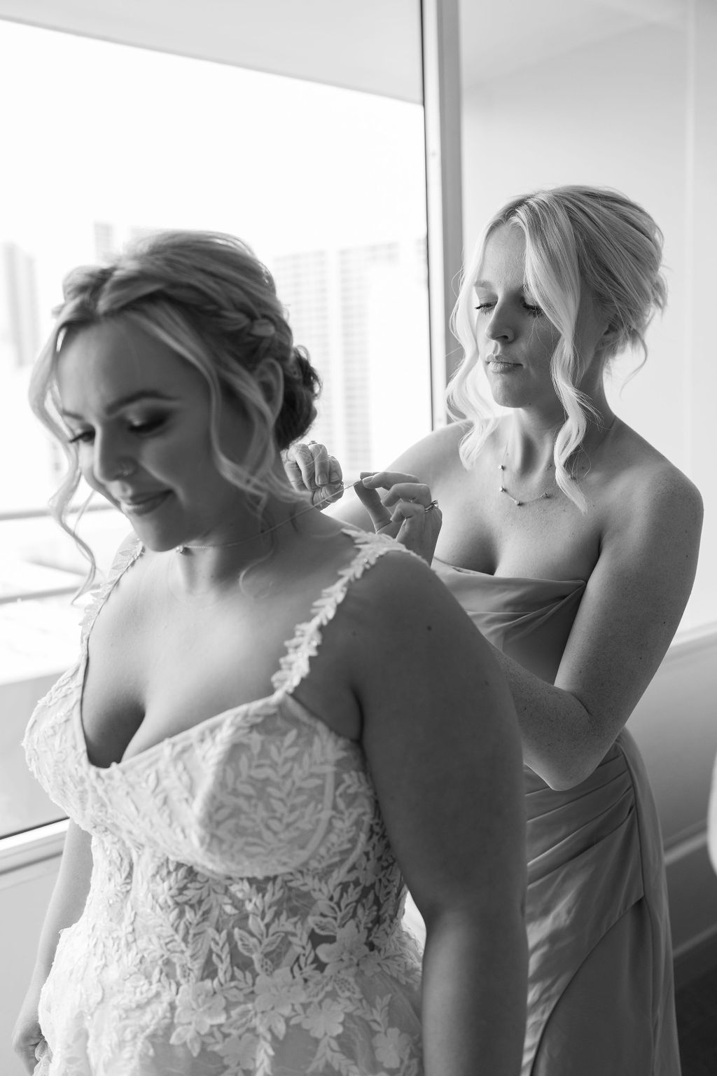 A woman in a wedding dress stands as another woman in a bridesmaid dress adjusts her strap in a bright room.