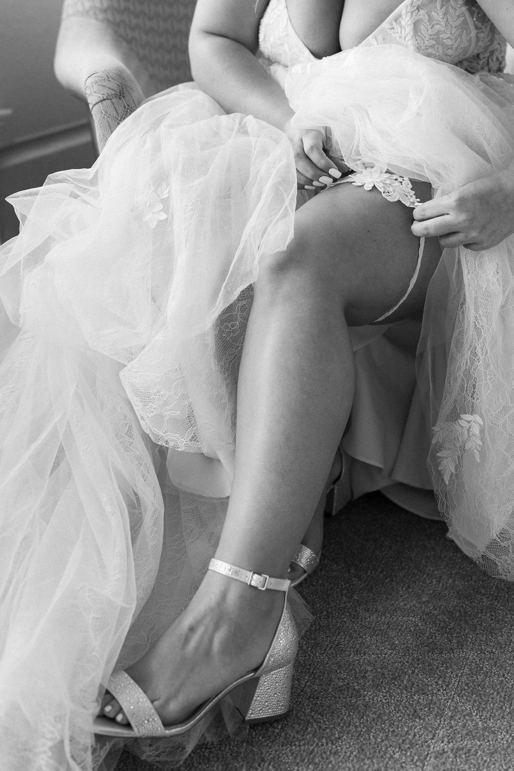 A woman in a wedding dress stands as another woman in a bridesmaid dress adjusts her strap in a bright room.