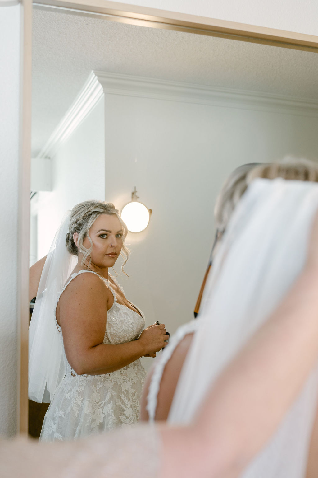 A bride in a white gown and veil looks at herself in a mirror, with her reflection clearly visible.