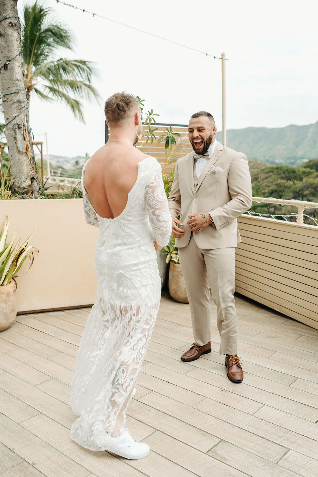  Two people stand on a deck outdoors; one wears a white lace dress and sneakers, the other wears a tan suit and brown shoes. They face each other, appearing to smile.