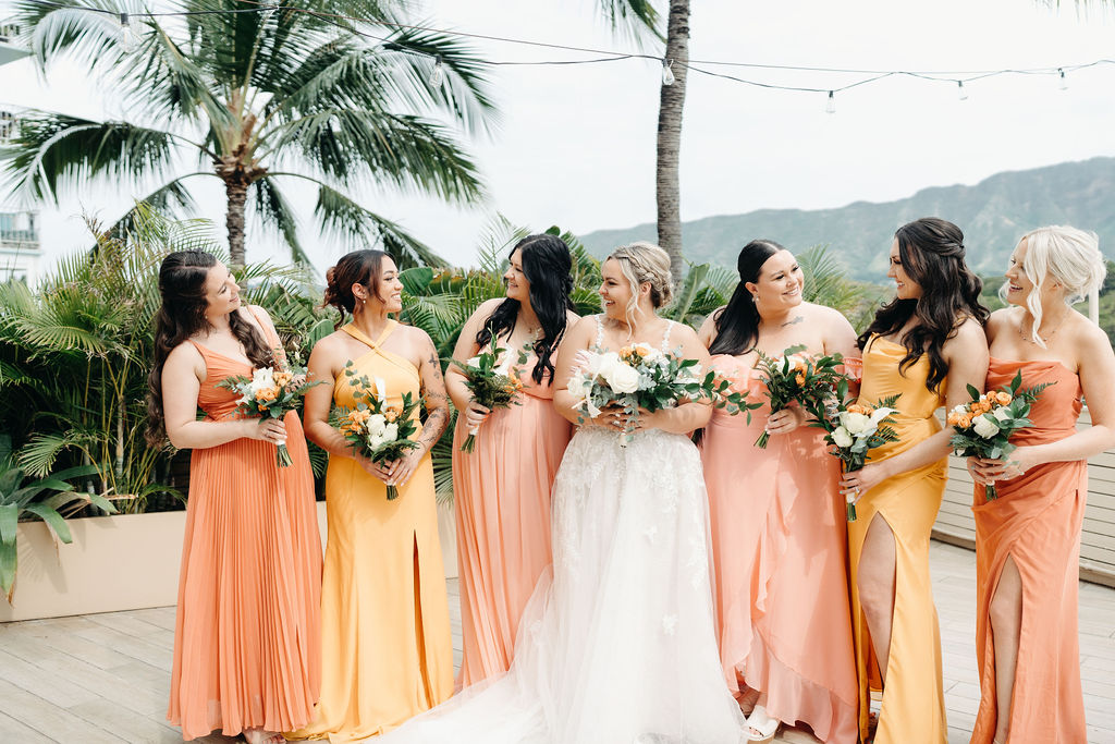 A bride in a white dress stands on a deck as four bridesmaids in orange dresses adjust her gown. Tropical plants and mountains are visible in the background for a wedding at queen kapiolani