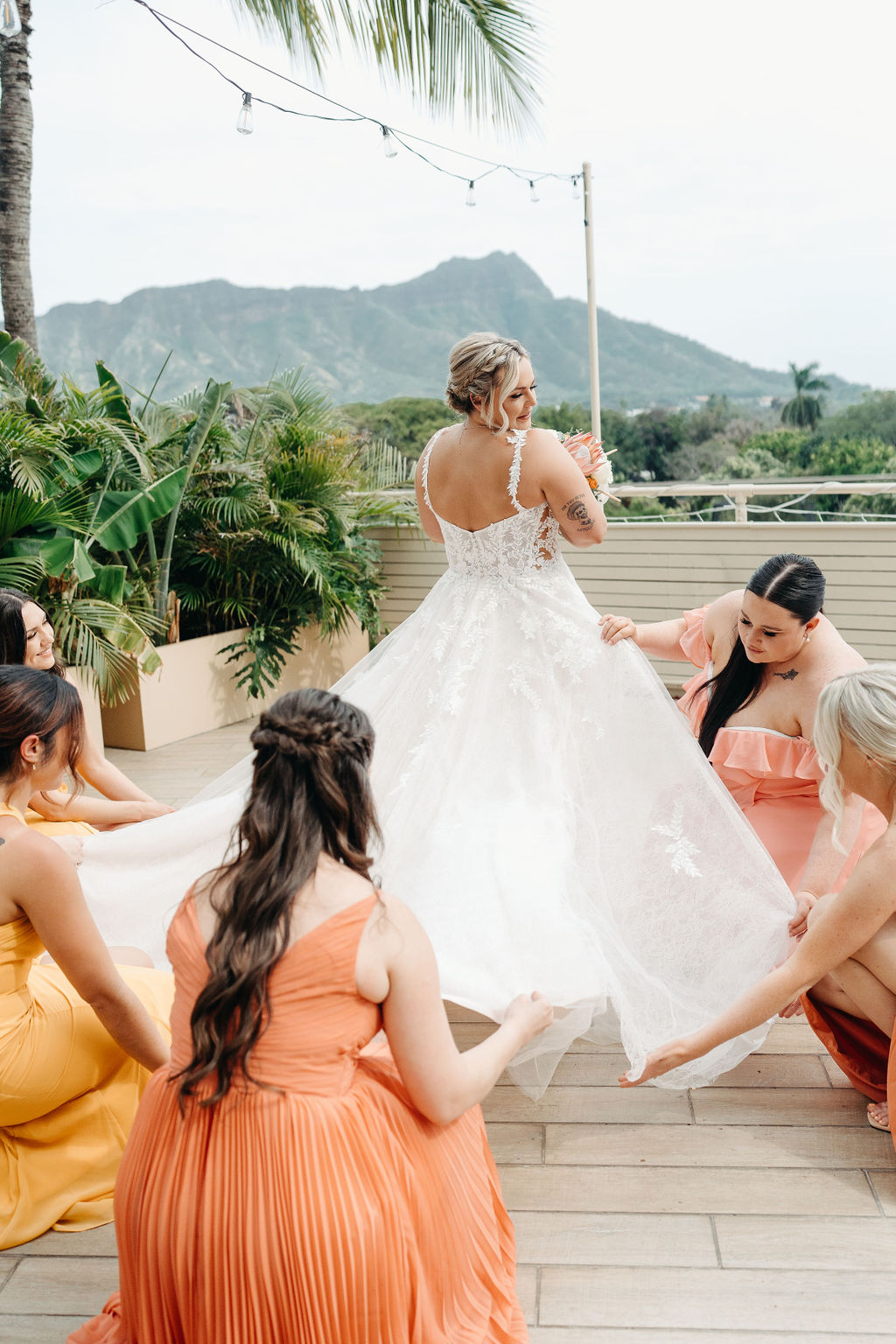 A bride in a white dress stands on a deck as four bridesmaids in orange dresses adjust her gown. Tropical plants and mountains are visible in the background for a wedding at queen kapiolani