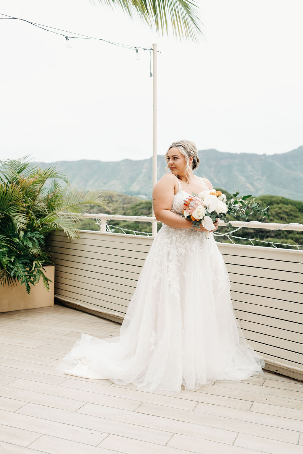 A bride in a white wedding dress holding a bouquet stands on an outdoor terrace with mountains and greenery in the background.