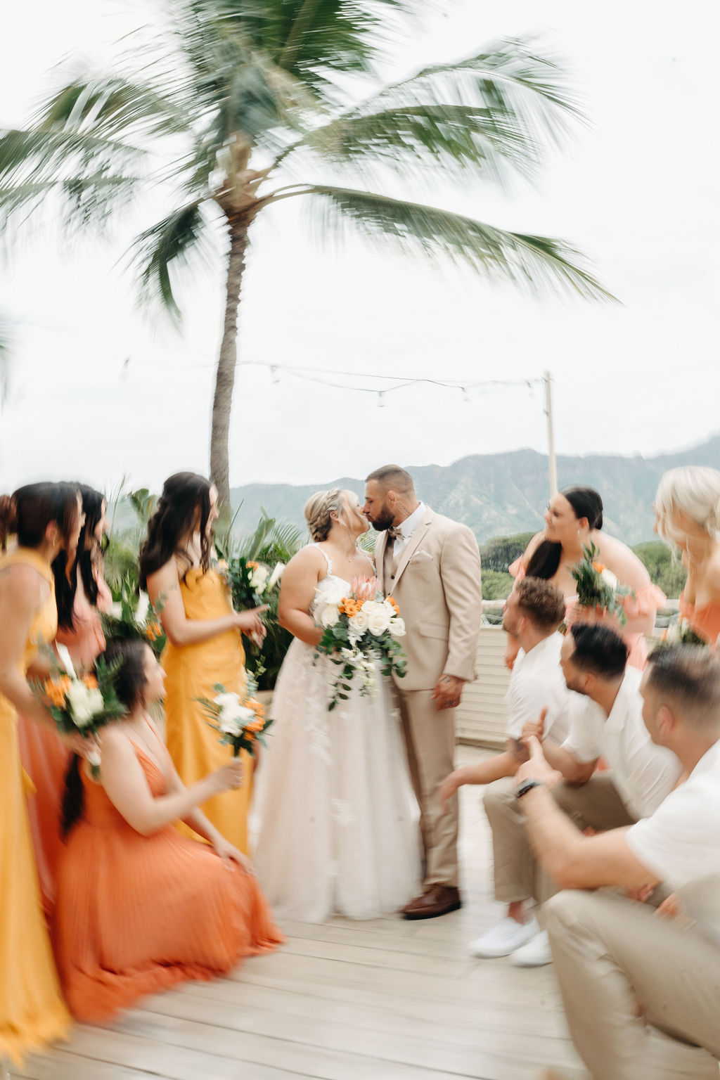 A bride in a white dress stands on a deck as four bridesmaids in orange dresses adjust her gown. Tropical plants and mountains are visible in the background for a wedding at queen kapiolani