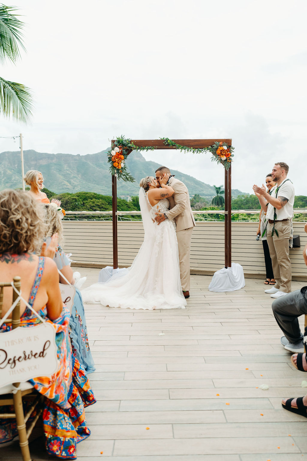 A bride and groom stand under a decorated wooden arch outdoors, facing each other as the bride reads from a paper. Mountains and greenery are visible in the background for a wedding at queen kapiolani hotel