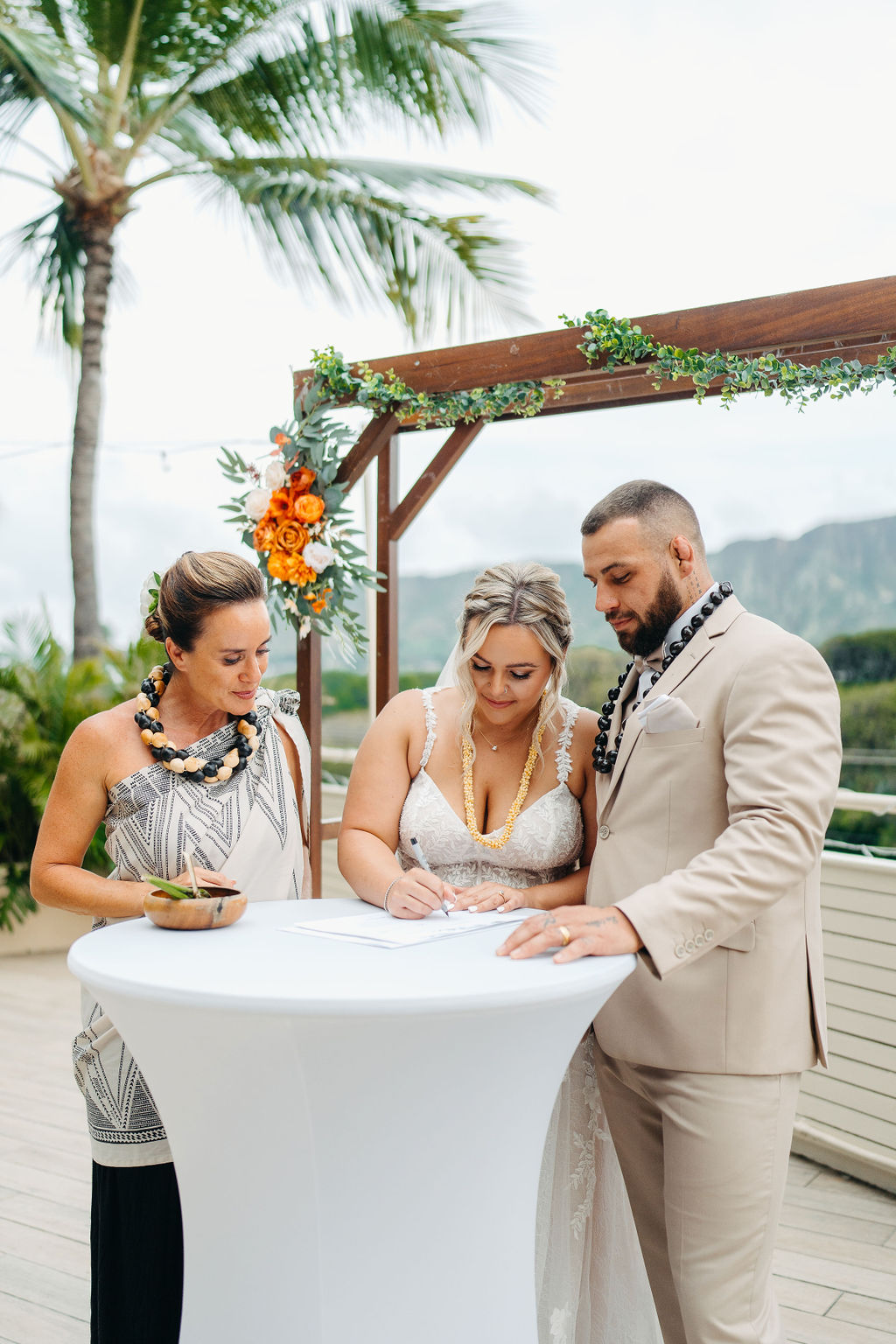 A bride and groom stand under a decorated wooden arch outdoors, facing each other as the bride reads from a paper. Mountains and greenery are visible in the background for a wedding at queen kapiolani hotel