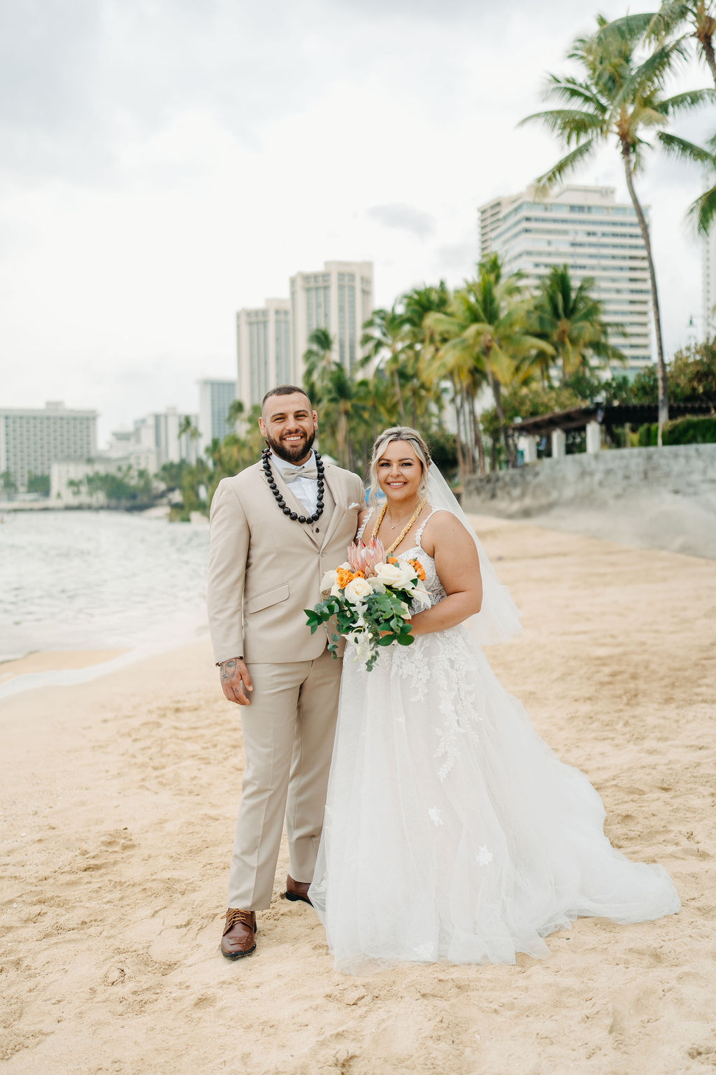 A bride and groom in wedding attire stand on a beach, touching foreheads and holding a bouquet of white and orange flowers for a wedding at Queen Kapiolani Hotel