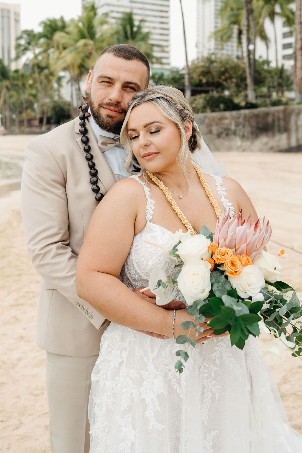 A bride and groom in wedding attire stand on a beach, touching foreheads and holding a bouquet of white and orange flowers for a wedding at Queen Kapiolani Hotel
