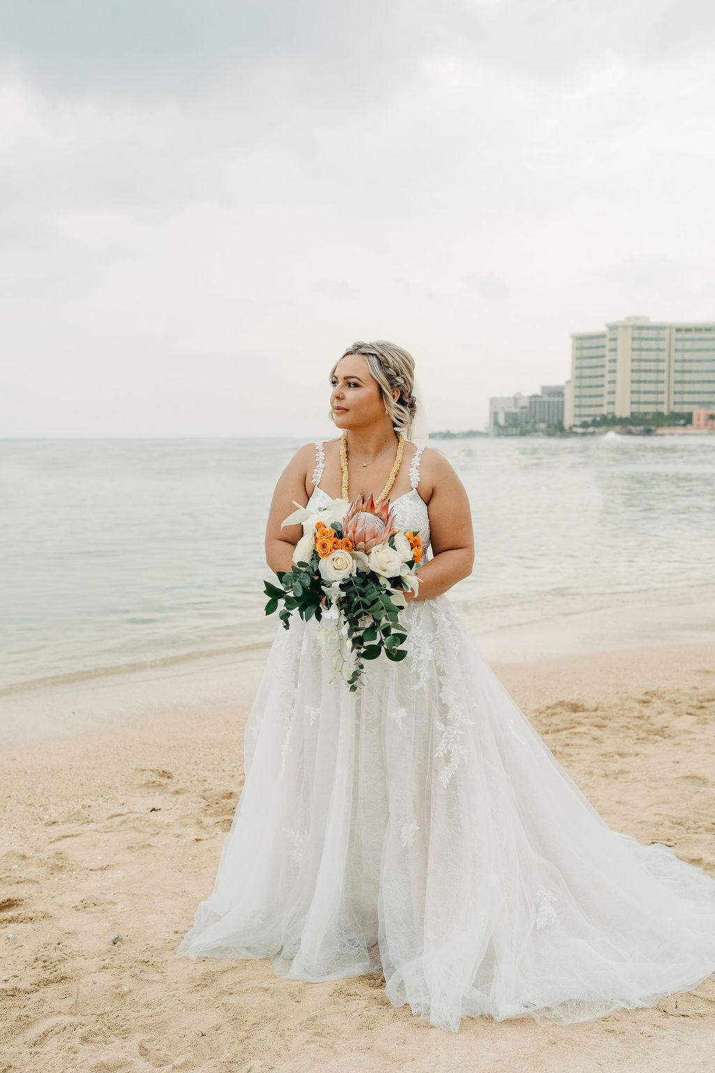 A bride and groom in wedding attire stand on a beach, touching foreheads and holding a bouquet of white and orange flowers for a wedding at Queen Kapiolani Hotel