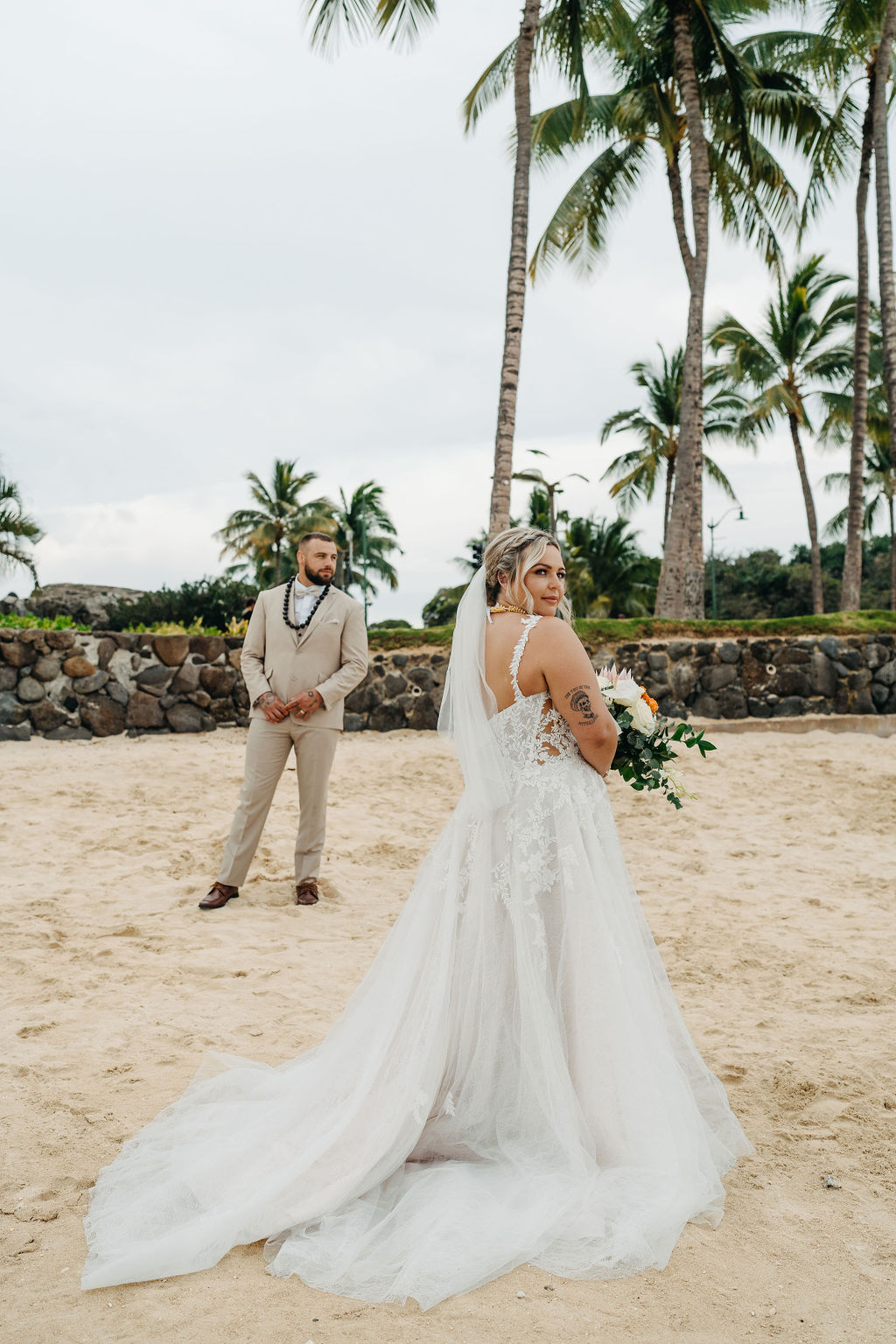 A bride and groom in wedding attire stand on a beach, touching foreheads and holding a bouquet of white and orange flowers for a wedding at Queen Kapiolani Hotel