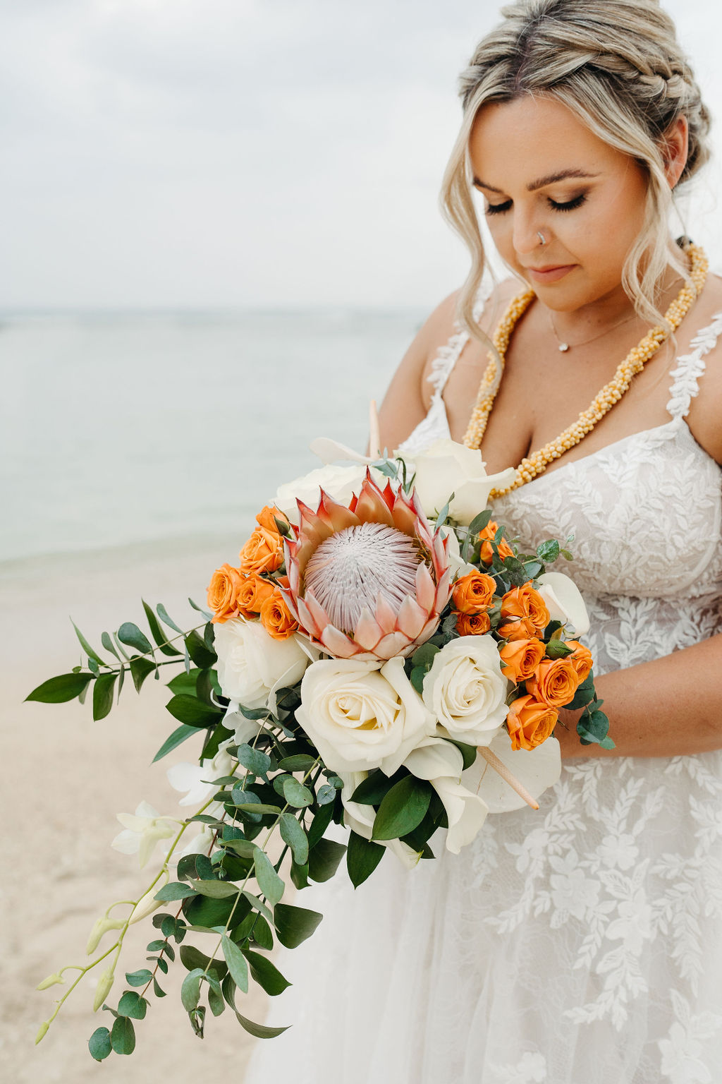 A bride and groom in wedding attire stand on a beach, touching foreheads and holding a bouquet of white and orange flowers for a wedding at Queen Kapiolani Hotel