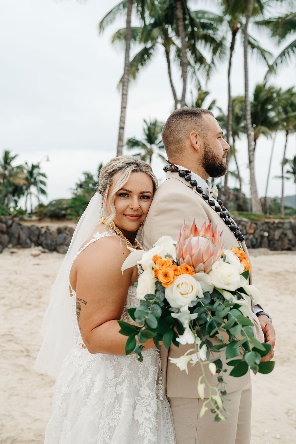 A bride and groom in wedding attire stand on a beach, touching foreheads and holding a bouquet of white and orange flowers for a wedding at Queen Kapiolani Hotel