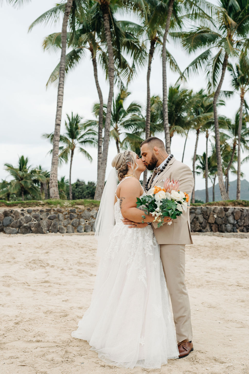 A bride and groom in wedding attire stand on a beach, touching foreheads and holding a bouquet of white and orange flowers for a wedding at Queen Kapiolani Hotel