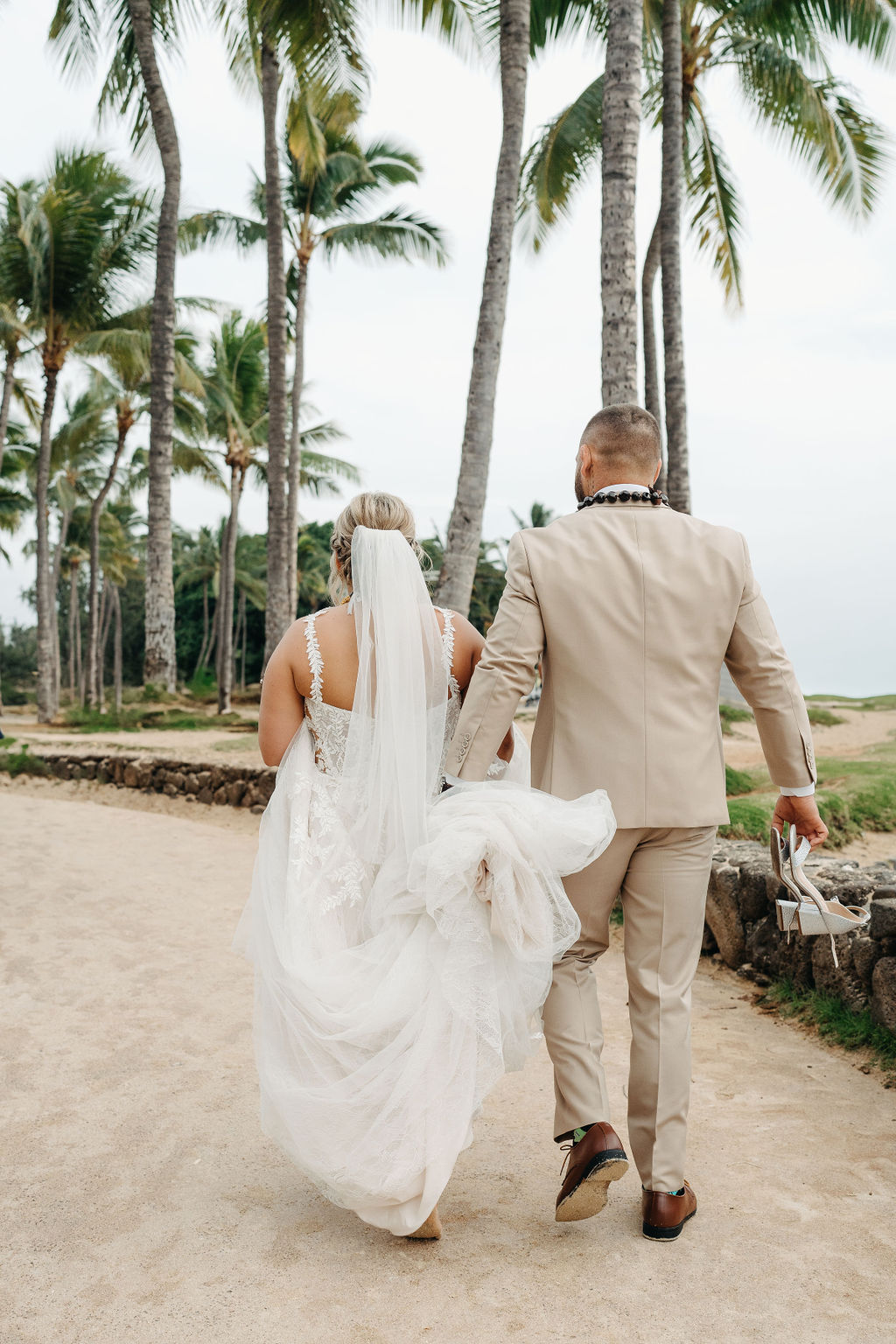A bride and groom in wedding attire stand on a beach, touching foreheads and holding a bouquet of white and orange flowers for a wedding at Queen Kapiolani Hotel