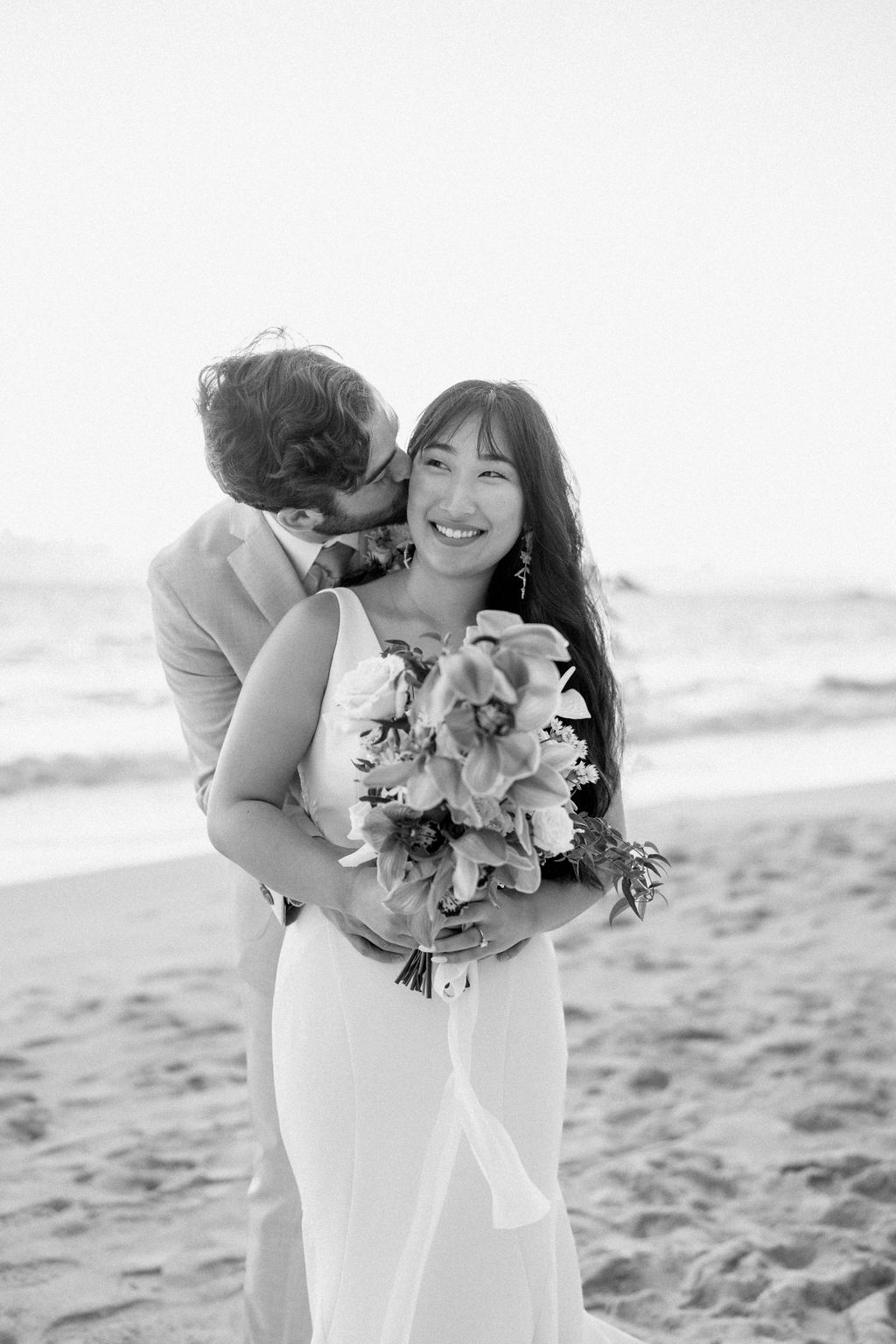 A couple in wedding attire embraces on a beach, with the ocean and sky in the background. The woman holds a bouquet and both appear happy for their Waimanalo Beach elopement
