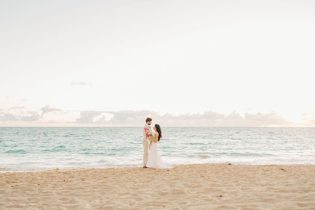 A couple in wedding attire embraces on a beach, with the ocean and sky in the background. The woman holds a bouquet and both appear happy for their Waimanalo Beach elopement