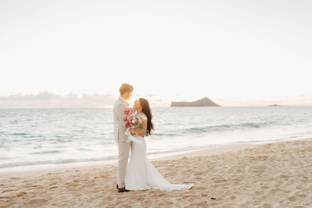 A couple in wedding attire embraces on a beach, with the ocean and sky in the background. The woman holds a bouquet and both appear happy for their Waimanalo Beach elopement
