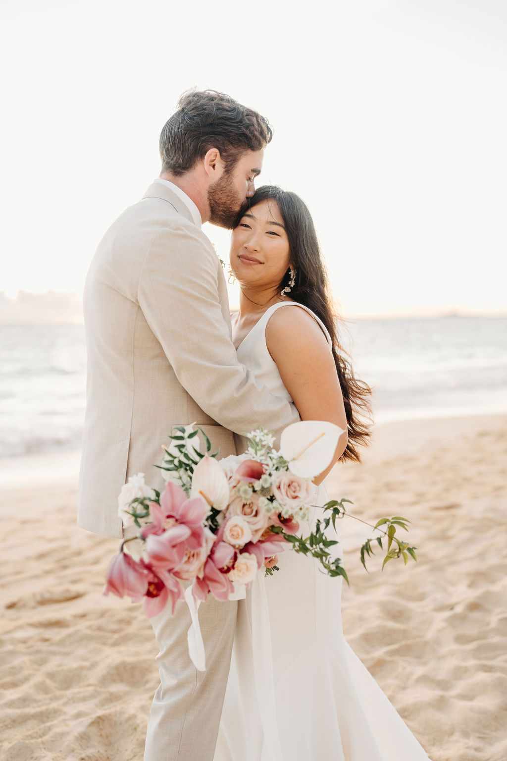 A couple in wedding attire embraces on a beach, with the ocean and sky in the background. The woman holds a bouquet and both appear happy for their Waimanalo Beach elopement