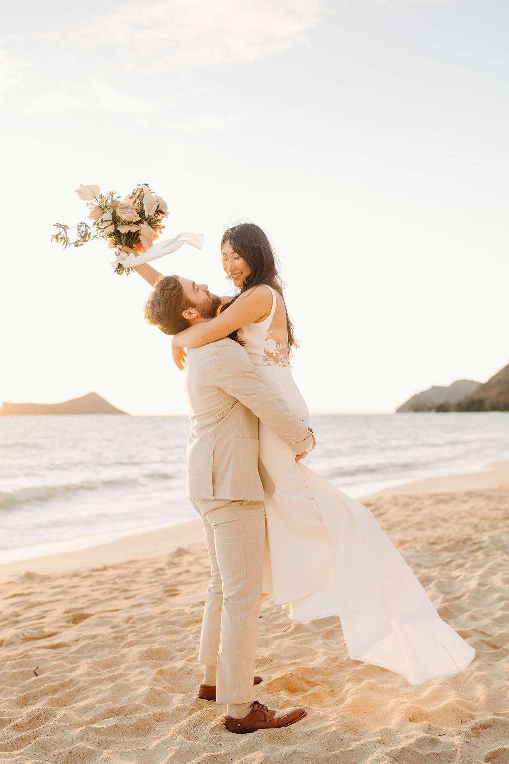A couple in wedding attire embraces on a beach, with the ocean and sky in the background. The woman holds a bouquet and both appear happy for their Waimanalo Beach elopement