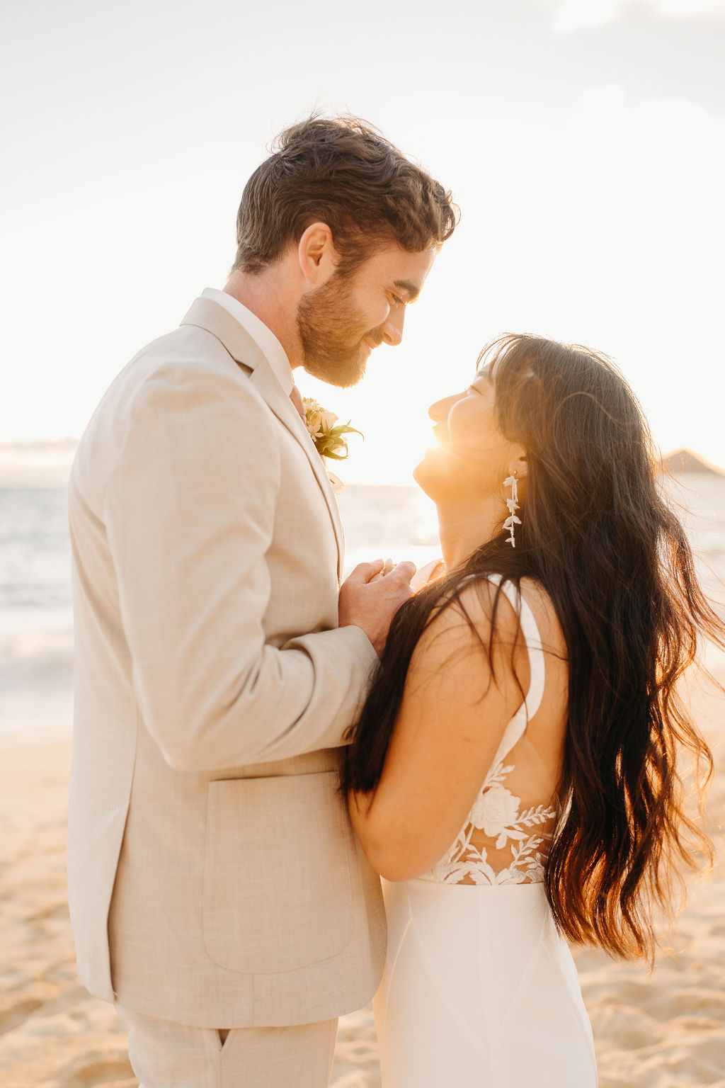 A couple in wedding attire embraces on a beach, with the ocean and sky in the background. The woman holds a bouquet and both appear happy for their Waimanalo Beach elopement