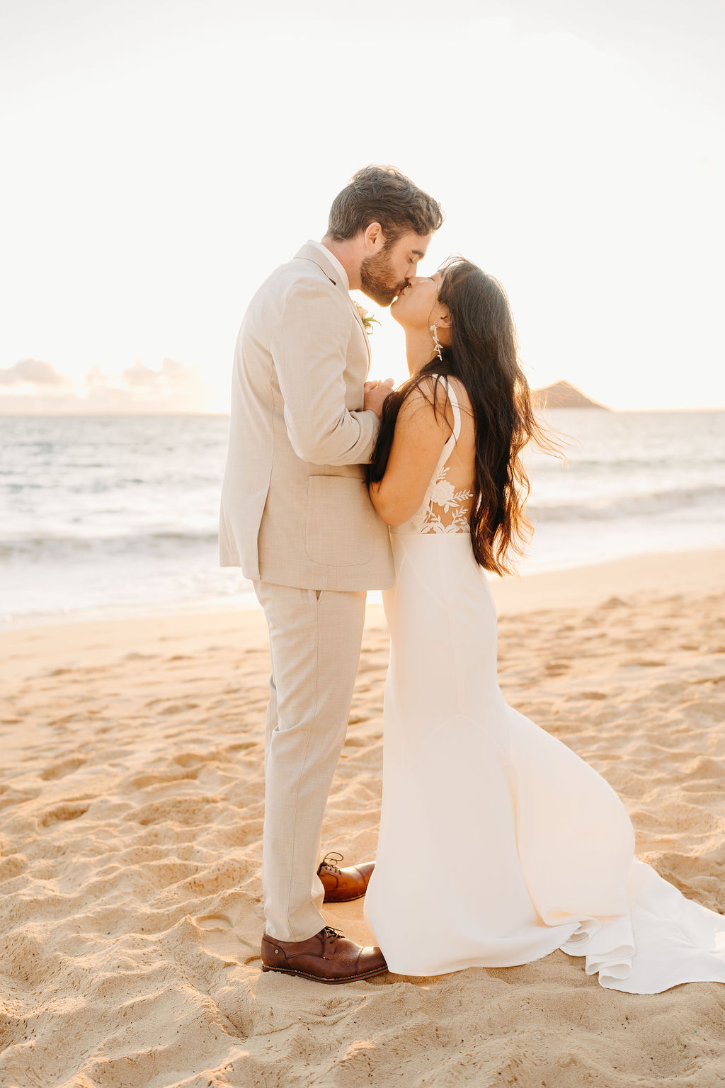 A couple in wedding attire embraces on a beach, with the ocean and sky in the background. The woman holds a bouquet and both appear happy for their Waimanalo Beach elopement
