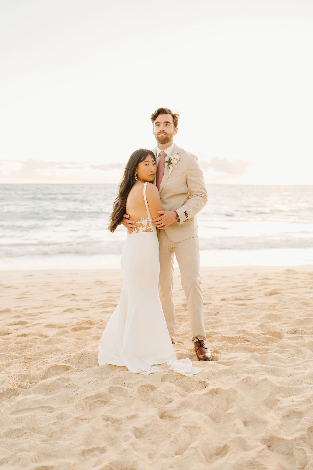 A couple in wedding attire embraces on a beach, with the ocean and sky in the background. The woman holds a bouquet and both appear happy for their Waimanalo Beach elopement