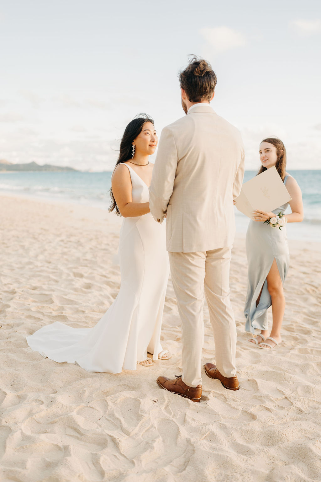 A bride and groom stand facing each other with an officiant between them, ocean waves and a bright sky in the background.