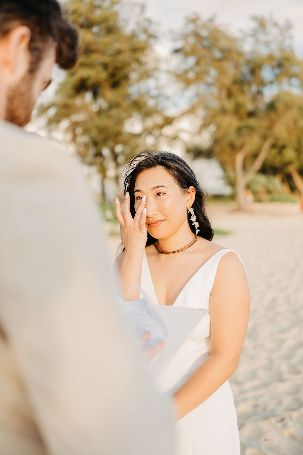 A bride and groom stand facing each other with an officiant between them, ocean waves and a bright sky in the background.