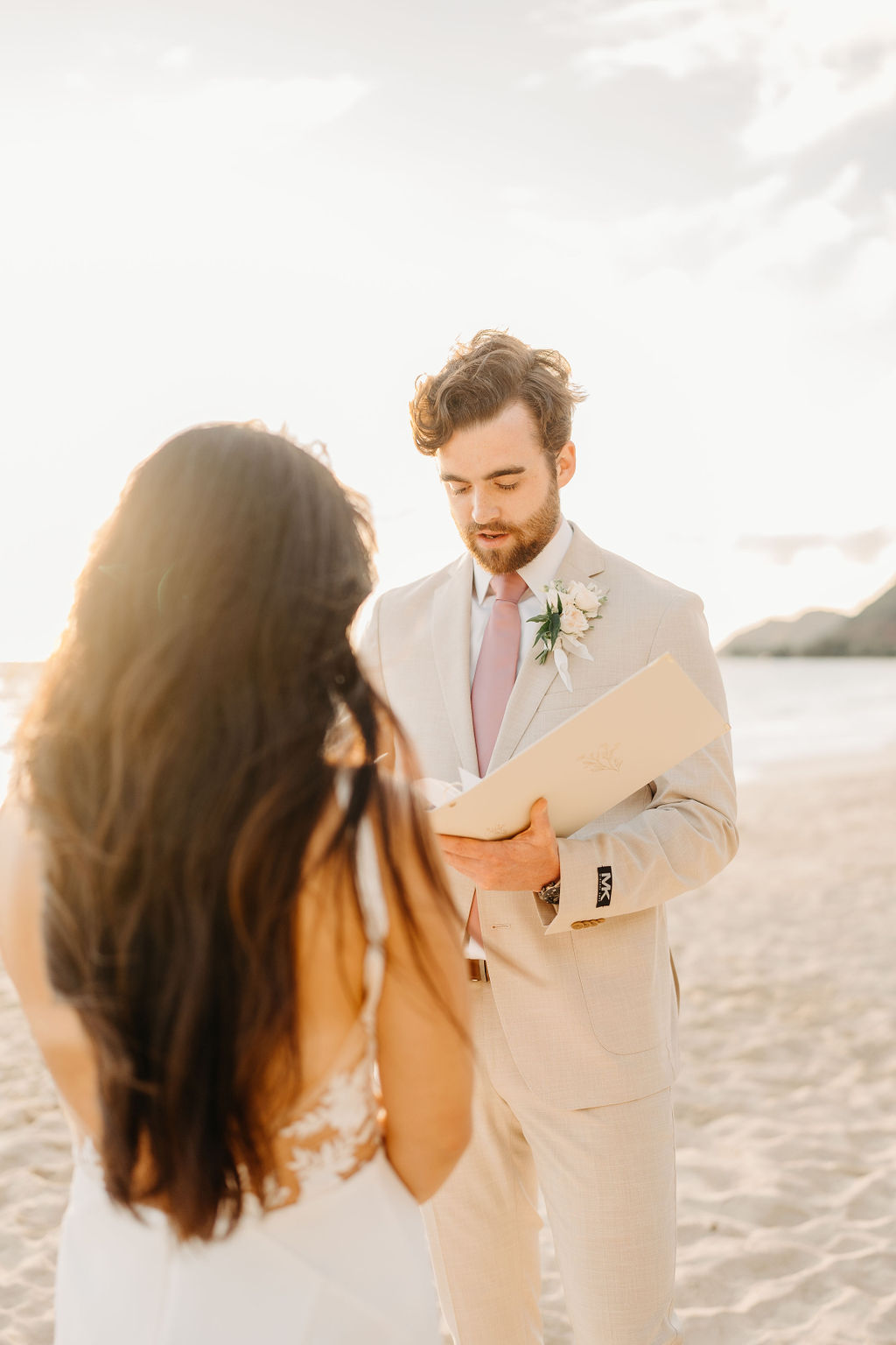 A bride and groom stand facing each other with an officiant between them, ocean waves and a bright sky in the background.