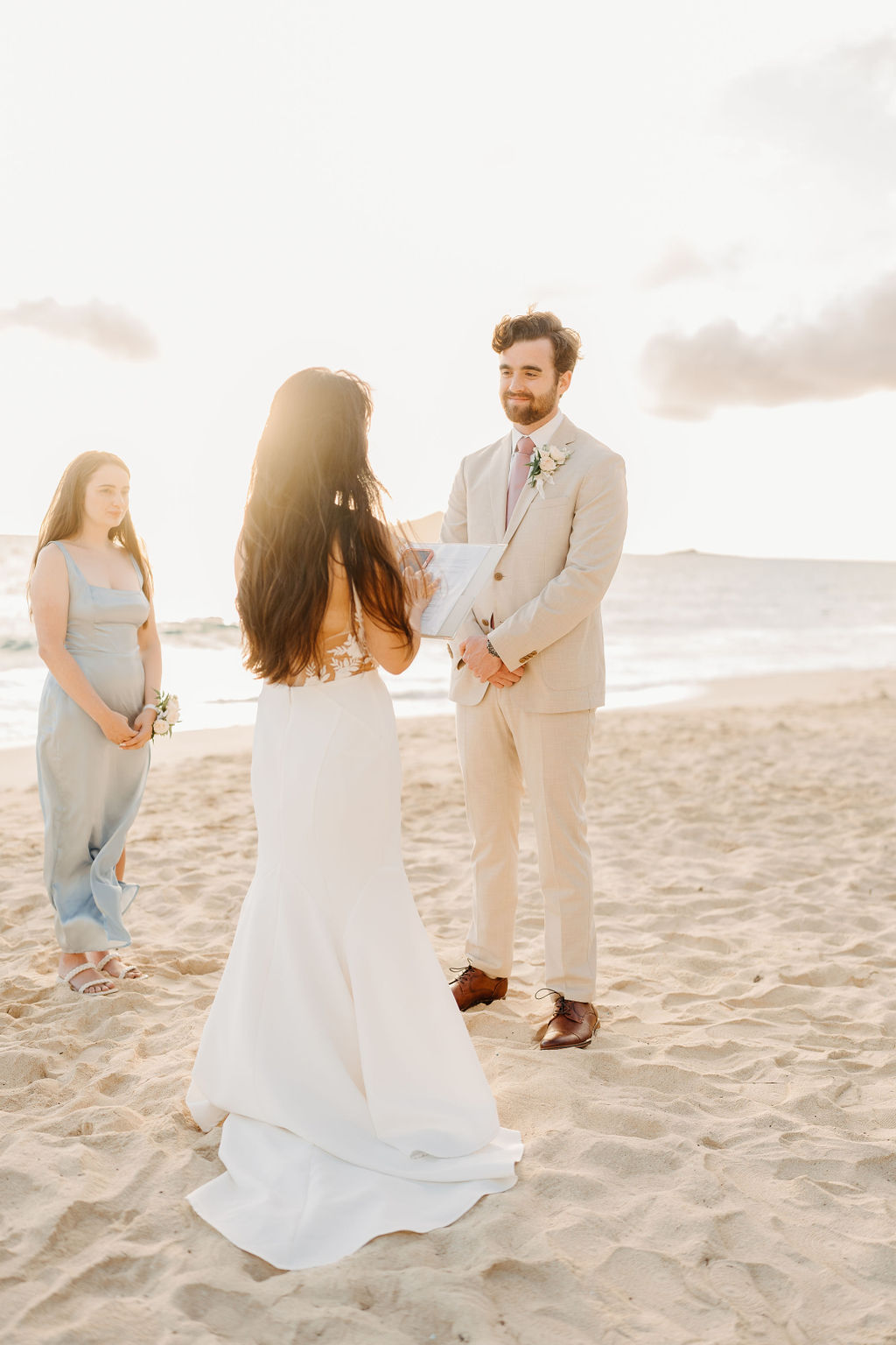 A bride and groom stand facing each other with an officiant between them, ocean waves and a bright sky in the background.