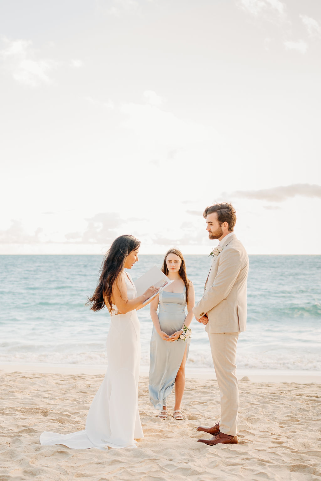 A bride and groom stand facing each other with an officiant between them, ocean waves and a bright sky in the background.