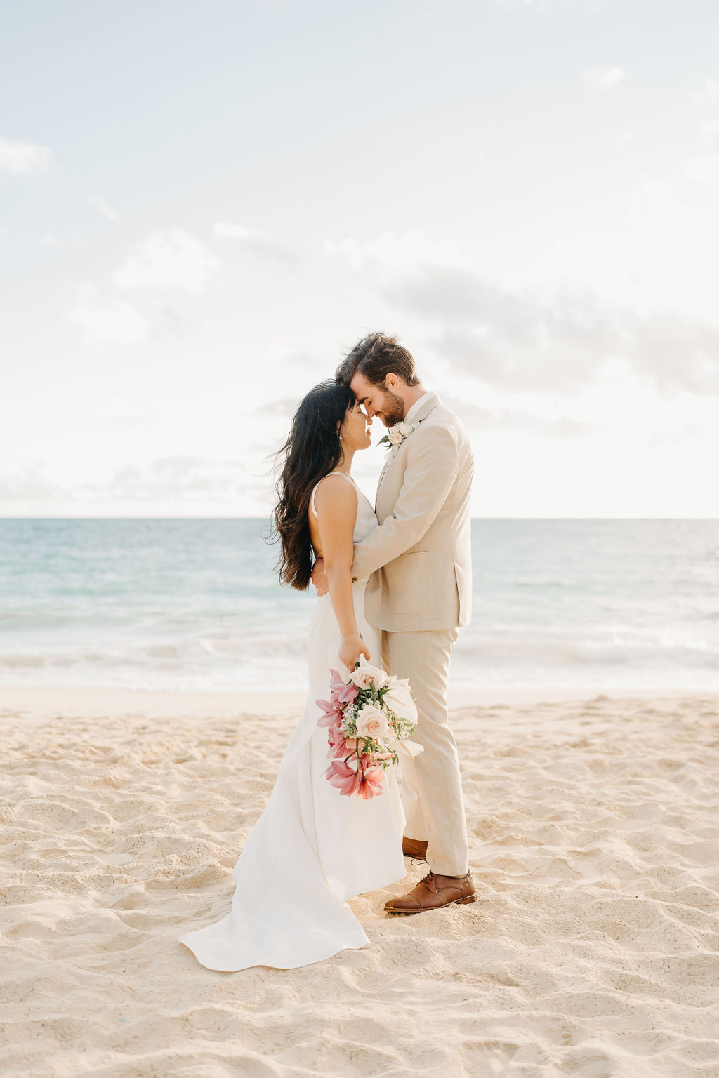 A couple in wedding attire embraces with the ocean and sky in the background. The woman holds a bouquet