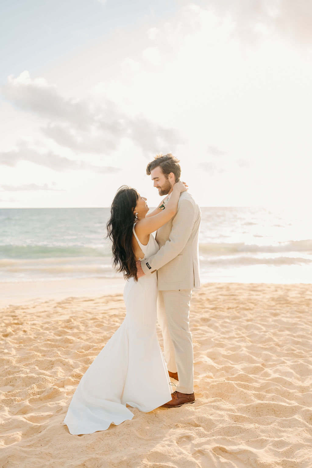 A couple in wedding attire embraces with the ocean and sky in the background. The woman holds a bouquet