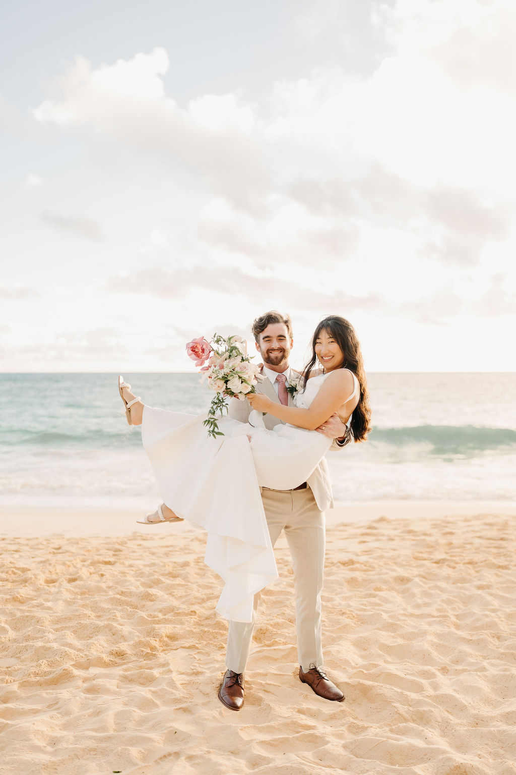 A couple in wedding attire embraces with the ocean and sky in the background. The woman holds a bouquet