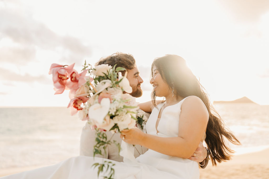 A couple in wedding attire embraces  with the ocean and sky in the background. The woman holds a bouquet